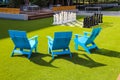 Three blue lawn chairs near a giant chess set on lush green grass in the park surrounded by lush green trees and buildings