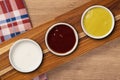 Three bowls with sauces, ketchup, mustard, white, on a wooden background. top view