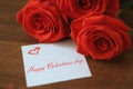 Three bright red roses and a small sheet of paper with the inscription!`Happy Valentines day!` on a wooden brown background closeu