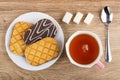 Checkered cookies in chocolate on plate, sugar cubes, teaspoon, cup of tea on table. Top view