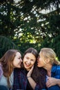 Three cheerful girls whisper and gossip against green foliage in the park. Women joke and laugh, vertical image
