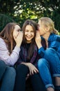 Three cheerful girls whisper and gossip against green foliage in the park. Women joke and laugh, vertical image