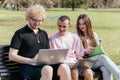 Three cheerful students study and work together outdoors on a sunny day, using laptops and notebooks while sitting on a Royalty Free Stock Photo