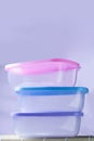 three clear plastic boxes with colored lids, stacked on top of each other, all three empty inside. Behind it is a blurred purple