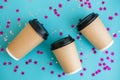 Three coffee cardboard craft cups lie on blue background with confetti. Mock-up. Top view. Flat lay.