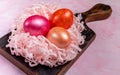 Three colored eggs in nest from pink paper shreds on dark wooden kitchen board on pink textured background. Close-up. Royalty Free Stock Photo