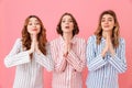 Three concentrated young girls 20s wearing colorful striped pajamas praying keeping palms together and wishing good luck during s