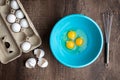 Three cracked eggs in blue mixing bowl, eggshells, whole eggs in cardboard carton, metal whisk, wood table