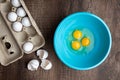 Three cracked eggs in blue mixing bowl, eggshells, whole eggs in cardboard carton, wood table