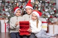 Three cute happy excited kids, boys and a girl in santa hats exchange surprise gifts next to the christmas tree at home Royalty Free Stock Photo