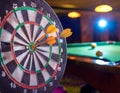 Three darts in a dart board with a pool game in the background, games room