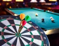Three darts in a dart board with a pool game in the background, games room