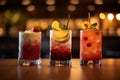 Three different colored cocktails on the bar counter with ice spread around and a shallow depth of field of the bar