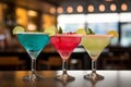 Three different colored cocktails on the bar counter with ice spread around and a shallow depth of field of the bar