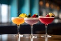 Three different colored cocktails on the bar counter with ice spread around and a shallow depth of field of the bar