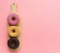 Three different donuts, chocolate donut, pink icing and without icing on a wooden cutting board on a pink background