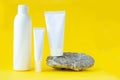 Three empty cosmetic packagings stand next to a natural stone on a yellow background. Copy space for text. Minimalism concept,