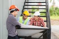 Three engineer wear helmet sit on a staircase and look at a construction plan or blueprint in white paper.