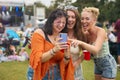 Three Female Friends Wearing Glitter Looking At Mobile Phone At Summer Music Festival Holding Drinks Royalty Free Stock Photo