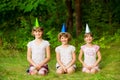 Three friendly children in festive cone caps, sit on grass, have fun together as celebrate birthday Royalty Free Stock Photo