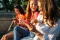 Three girl friends watching mobile smart phones and having fun sitting on bench in public park. Gen z, Teenagers devices Royalty Free Stock Photo