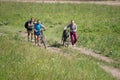 Three girls climb the rural road up the hill with bicycles, among the green grass