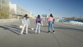 Three girls roller skate by harbor in sunlight, slomo from behind