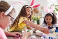 Three Girls Wearing Bunny Ears Sitting At Table Decorating Eggs For Easter At Home