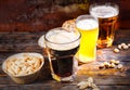 Three glasses with light, unfiltered and dark beer stand in a row near plates with snacks and scattered nuts on dark wooden desk