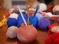 Three gold, green and white crochet hooks in a ceramic vase against a background of several multicolored balls of woolen thread on