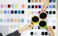 Three hands of woman hold 3 ceramic coffee cups and touch them together over working table with color sample catalog in the symbol Royalty Free Stock Photo