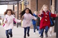 Three happy young school girls wearing coats and carrying schoolbags running in a walkway with their classmates outside their infa Royalty Free Stock Photo