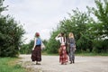 Three hippie women, wearing boho style clothes, holding guitar, walking on dirt road in countryside. Friends, traveling together