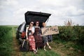 Three hippie women, wearing colorful boho style clothes, sitting on car trunk, holding Happy place sign, smiling, relaxing.