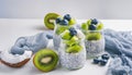 Three jars of chia seed pudding, topped with fresh kiwi slices and blueberries, beside a halved coconut and kiwi on a white