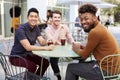 Three millennial male friends talking over cold brews outside a cafe in a city street turn to camera Royalty Free Stock Photo