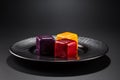 Three miniature cakes in the shape of a cube of different colors on a black plate.