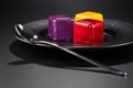 Three miniature cakes in the shape of a cube of different colors on a black plate.