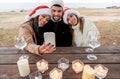 Three mixed race friends with lowered mask sitting on a wooden table outdoor in sea resort celebrating Christmas and New Year Royalty Free Stock Photo