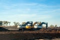 Three modern white and yellow excavators stand on excavated ground during road works on an intercity highway against a blue sky