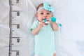 A three month baby girl in mint green clothes lying on a bed on which a measuring ruler for growth is drawn. Teething tool in