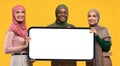 Three muslim women posing with modern smartphone with empty screen over yellow studio background Royalty Free Stock Photo