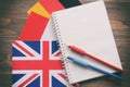 Three national flags, German, English, French, and notebook with blank sheets and two pencils on brown wooden surface. The concept