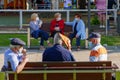 Three old men and three old women sat on opposite benches wearing face masks. One grandpa is protecting is head from the sun with