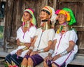 Three Padaung women in traditional dress and with metal rings around their neck are sitting next to each other in the village