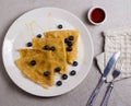 Three pancakes are on a white round plate, there are blueberries on the plate next to it