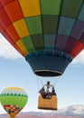 Three people lift off in the basket of a colorful hot air balloon, one woman looks back and takes a picture with her cell phone