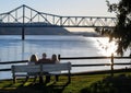 three people sitting on bench at sunset next to river and bridge Royalty Free Stock Photo