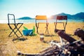 Three picnic chairs in the meadow field. Campfire on foreground and lake with mountain in background. Camping and traveling Royalty Free Stock Photo