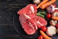 Three pieces of juicy raw beef with vegetables on a stone cutting board on a black wooden table background Royalty Free Stock Photo